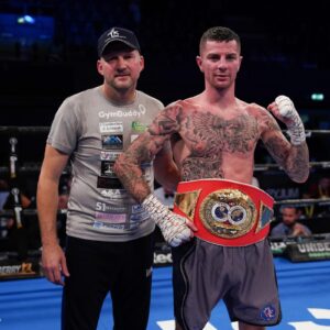 A boxer with tattoos and wrapped hands stands in a boxing ring wearing a championship belt, posing with his coach who is wearing a grey t-shirt and cap. Both look towards the camera after a bout.
