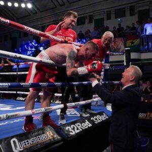 A boxer leans on the ropes in a ring, supported by his team. A man in a suit talks to him from outside the ring. The scene takes place in a crowded indoor arena with bright lighting.