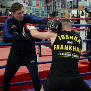 A boxing coach holds up pads while a boxer wearing a black Joshua Frankham Training Camp vest practises a punch inside a boxing ring. The gym walls are covered with posters.