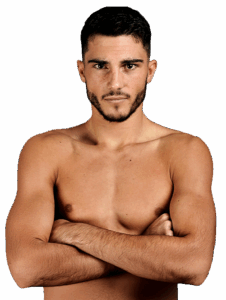 A bare-chested man with short dark hair and a beard stands with his arms folded, looking directly at the camera against a plain light background.