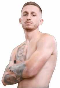 A bare-chested man with short hair and tattoos on his arms and chest stands with his arms folded, looking confidently at the camera against a plain light background.