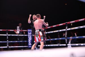 A boxer in red and white shorts stands in the ring with raised fists, while a referee cheers with both arms up. Spectators and boxing officials can be seen around the illuminated boxing ring.