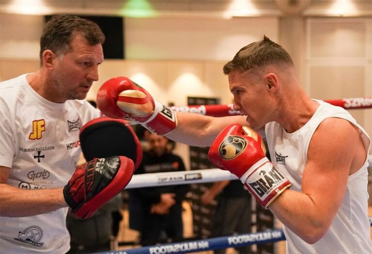 A boxer in a white vest throws a punch at focus pads held by a coach during a training session in a boxing ring. The coach wears a white shirt and red pads. Both appear focused and engaged.