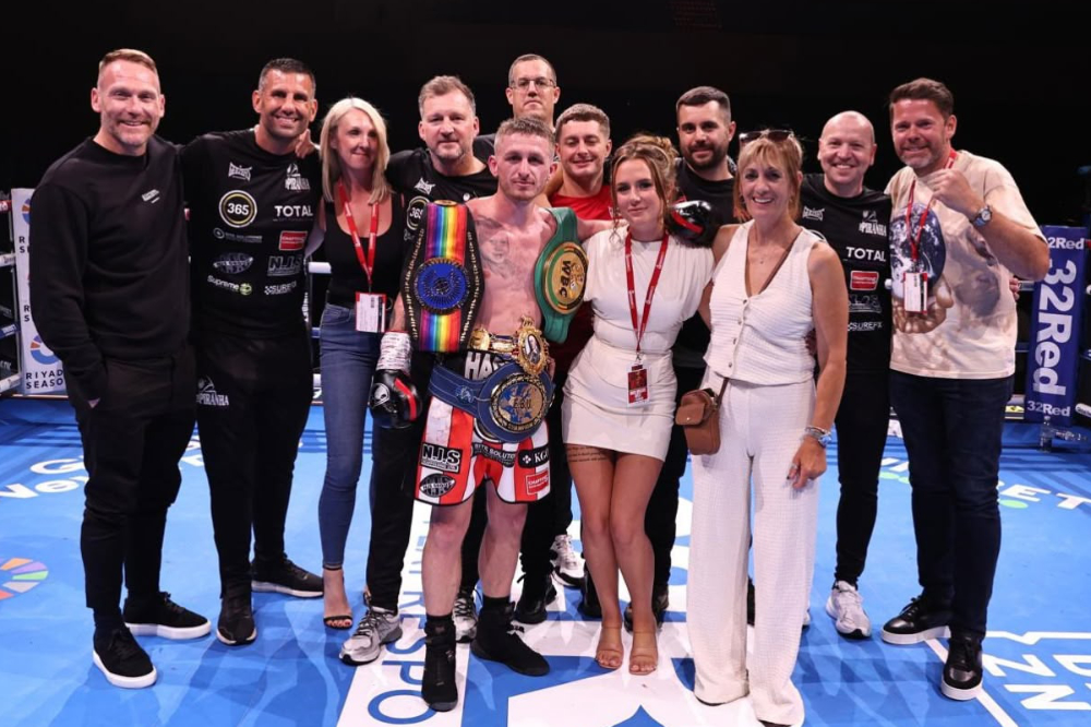 A victorious boxer stands in a ring holding championship belts, surrounded by smiling supporters and team members. Everyone is posing for a celebratory group photo after the bout.