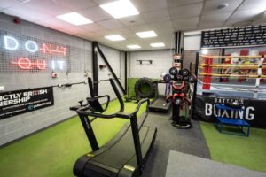Battens Boxing gym with a treadmill, boxing ring, punching equipment, neon “DON’T QUIT” sign, and green and grey flooring. The space is well-lit with motivational décor and training gear.
