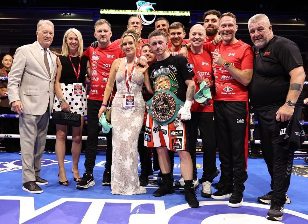 A boxer wearing a championship belt stands in a boxing ring, smiling and posing with his team and supporters after a fight. Everyone appears happy and celebratory.