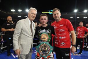 A boxer wearing a championship belt stands in a boxing ring between two men, one in a suit and one in a red shirt, after a fight. Bright arena lights shine in the background.