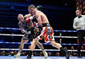 Two male boxers exchange powerful punches in a brightly lit boxing ring, with a referee watching closely and a crowd of spectators in the background.