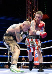 Two male boxers with tattoos fight in a boxing ring. One boxer throws a punch while the other ducks defensively. Spectators and bright lights are visible in the background.