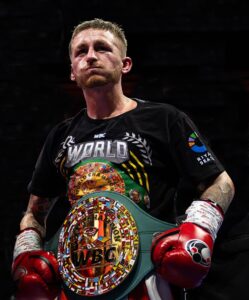 A male boxer wearing red gloves and a black T-shirt stands confidently, displaying a colourful WBC championship belt around his waist.